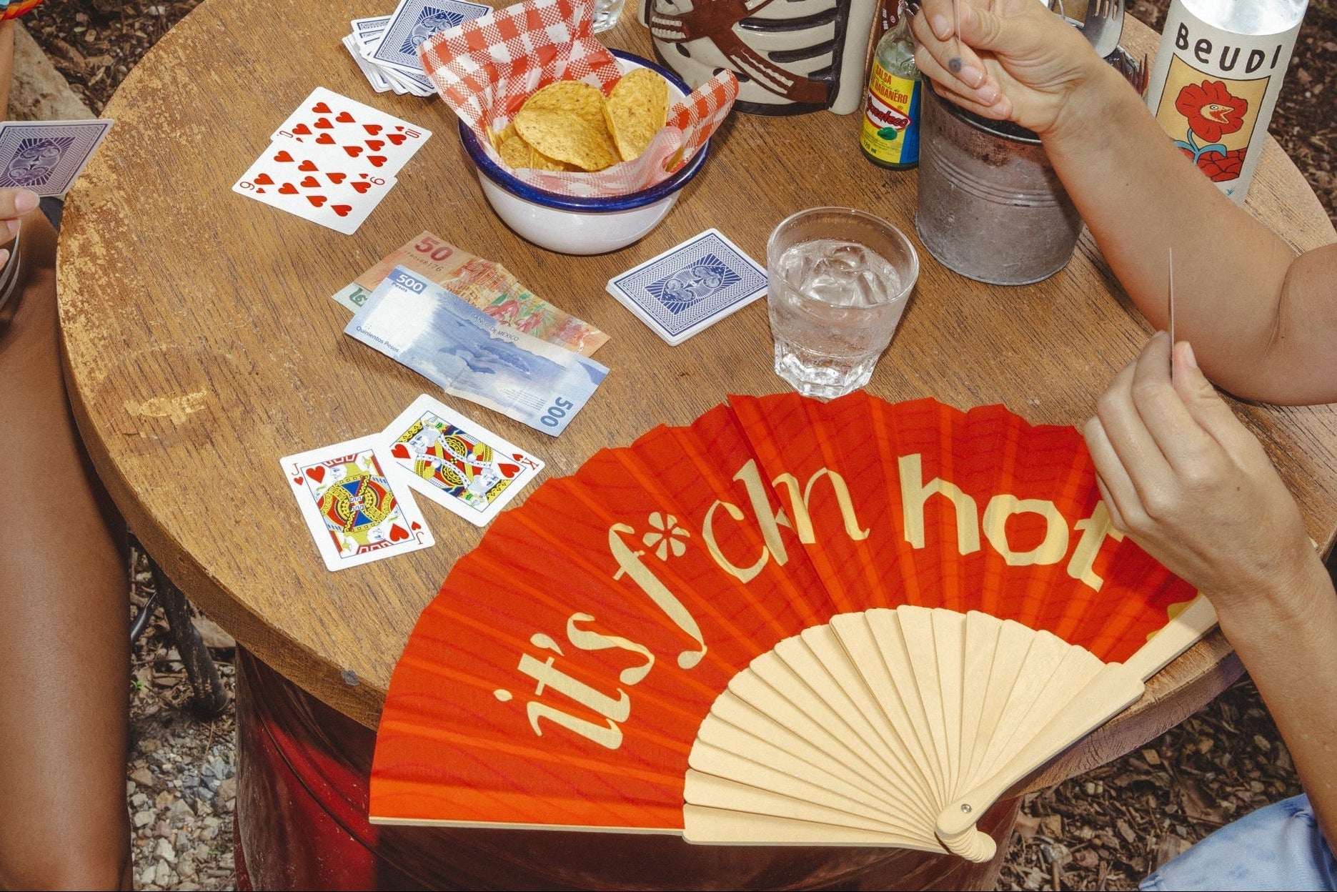 People playing cards around a table with snacks and drinks, including a 'it's so hot' fan.