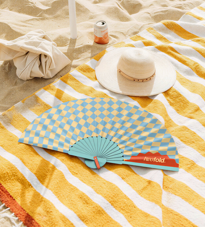 Blue and yellow foldable hand fan by Twofold on a beach towel, a stylish handheld fan for Australian summer heat, festivals and beach outings.
